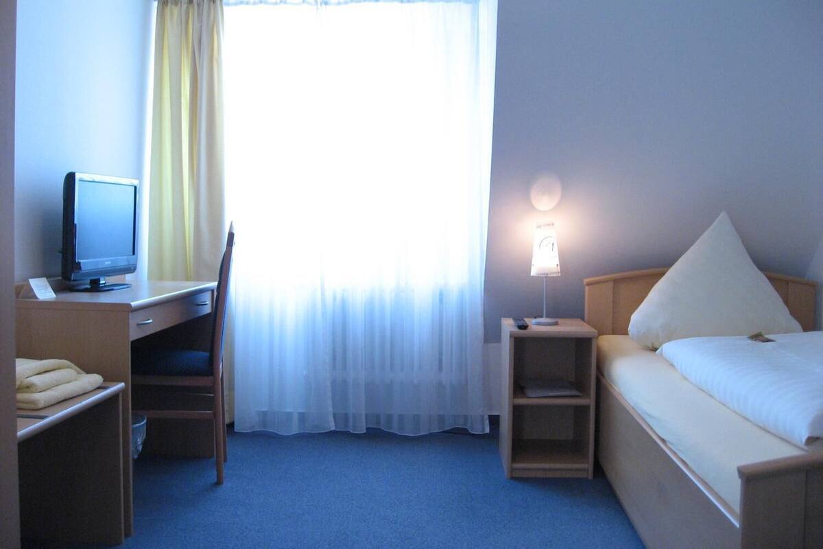 A hotel room with a bed, a desk with a computer, a bedside table with a lamp, and a window with curtains.