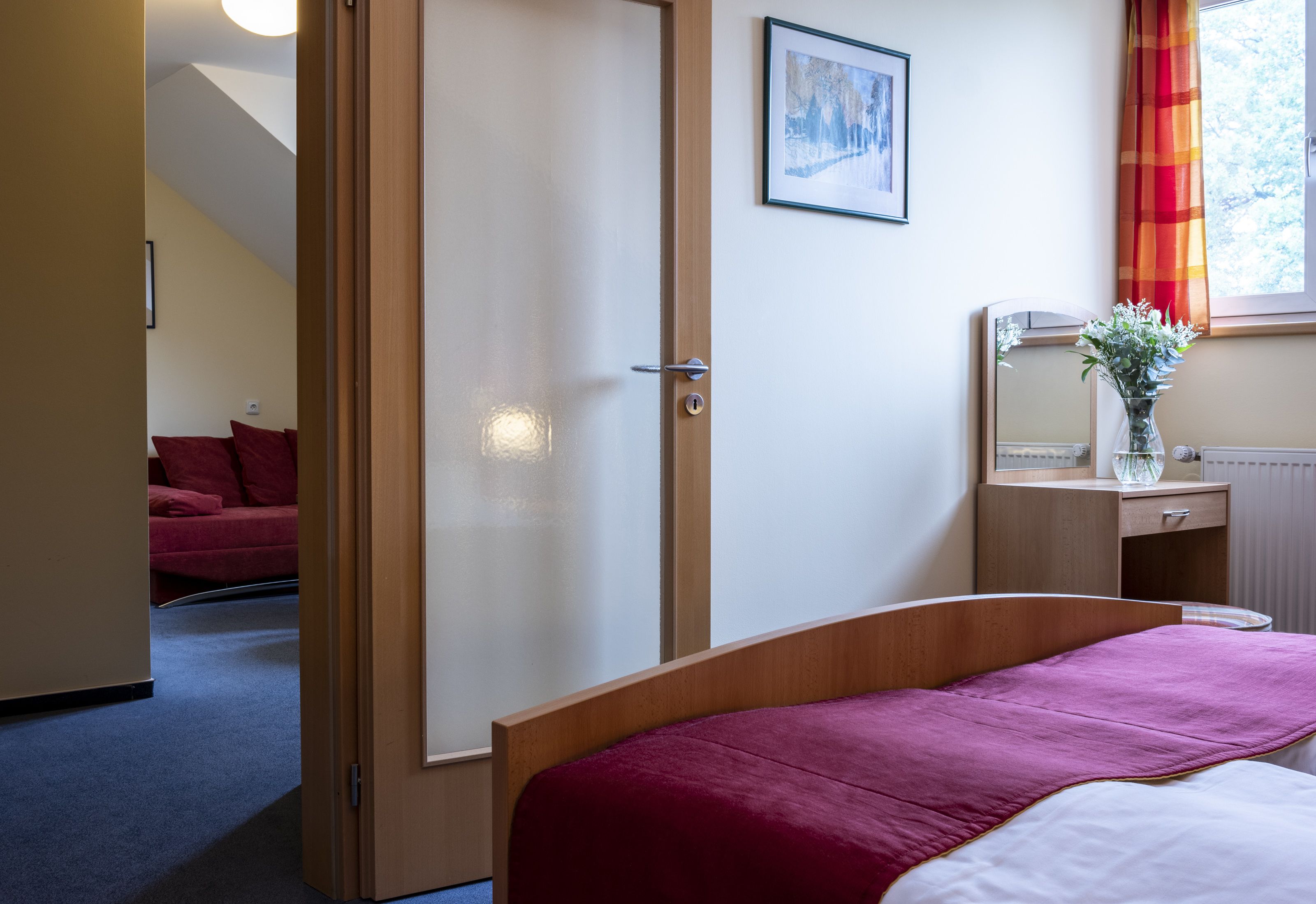 Ein Hotelzimmer mit Bett, einem Holzschrank, einem Nachttisch mit Vase, einem Fenster mit Vorhängen und einem roten Sofa im angrenzenden Raum.