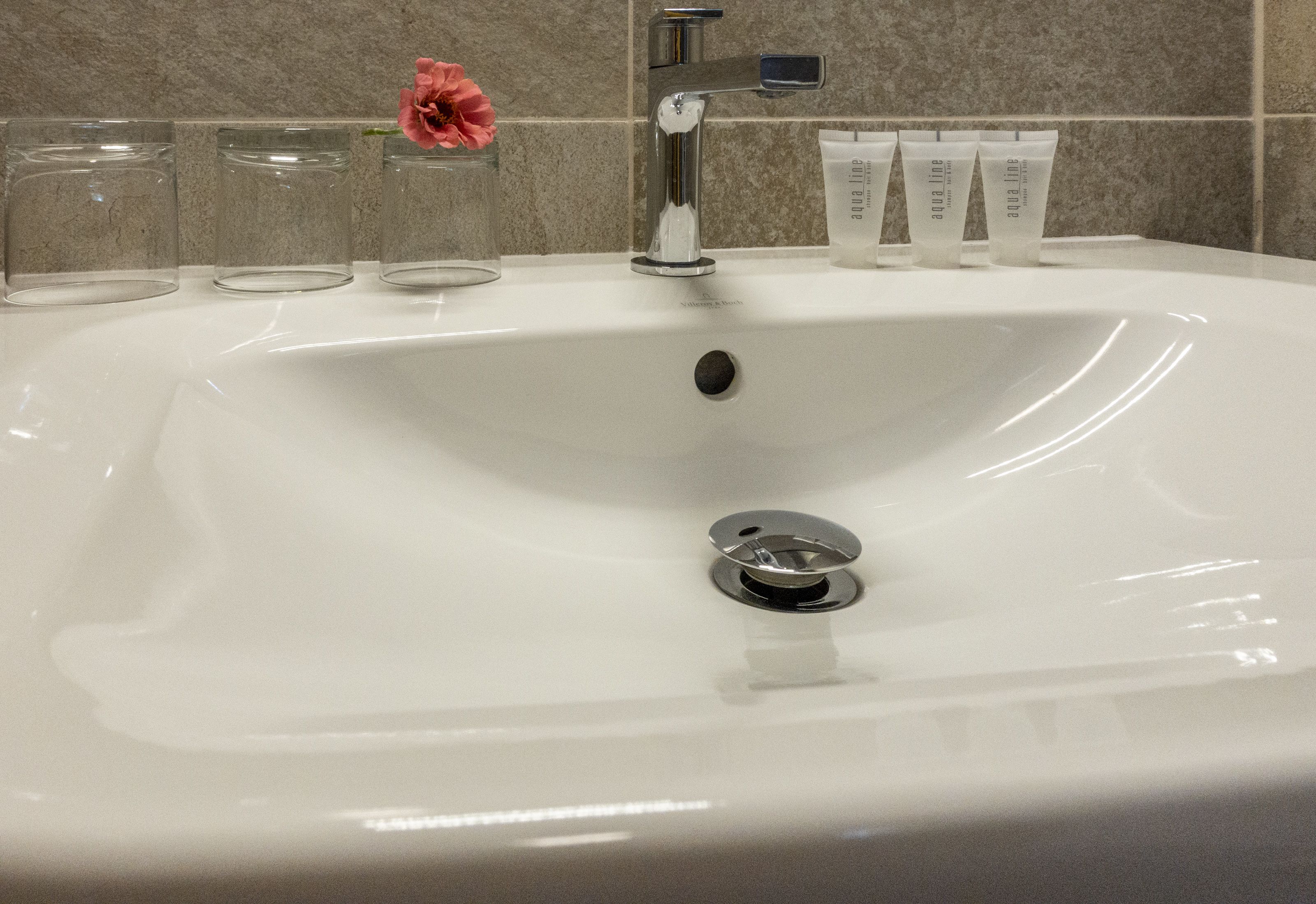 A bathroom sink with a chrome faucet, three clear glasses, and three tubes of Aquiline skincare products.