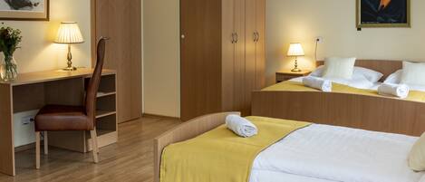 A hotel room with a wooden desk, a chair, a bed with a yellow blanket, and a nightstand with a lamp.