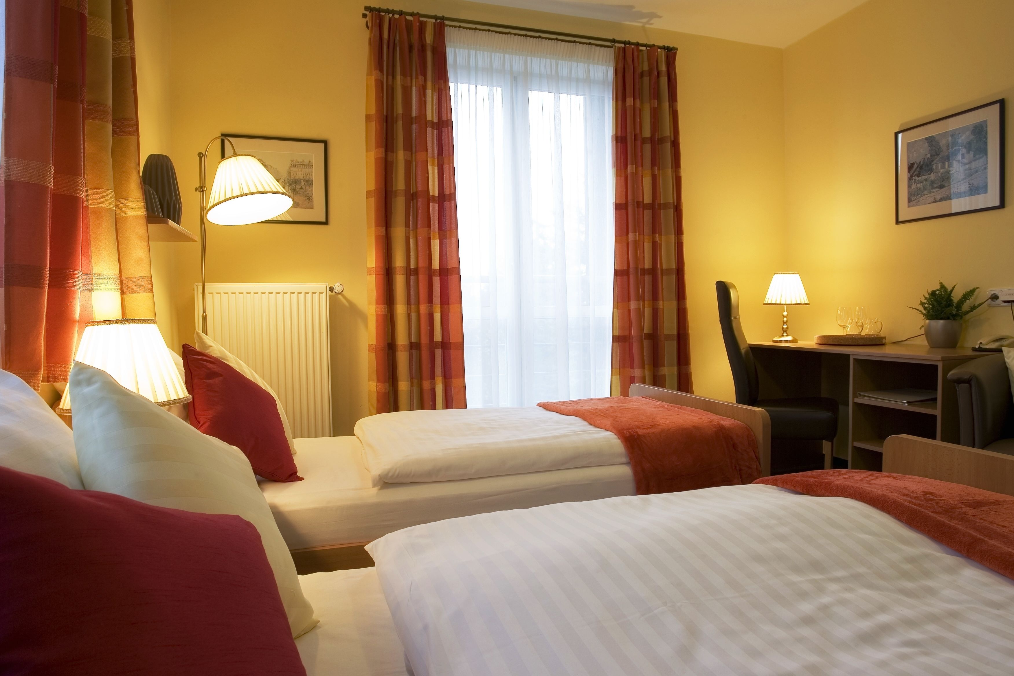 A hotel room with two beds, a desk, a chair, a radiator, and a window with curtains.