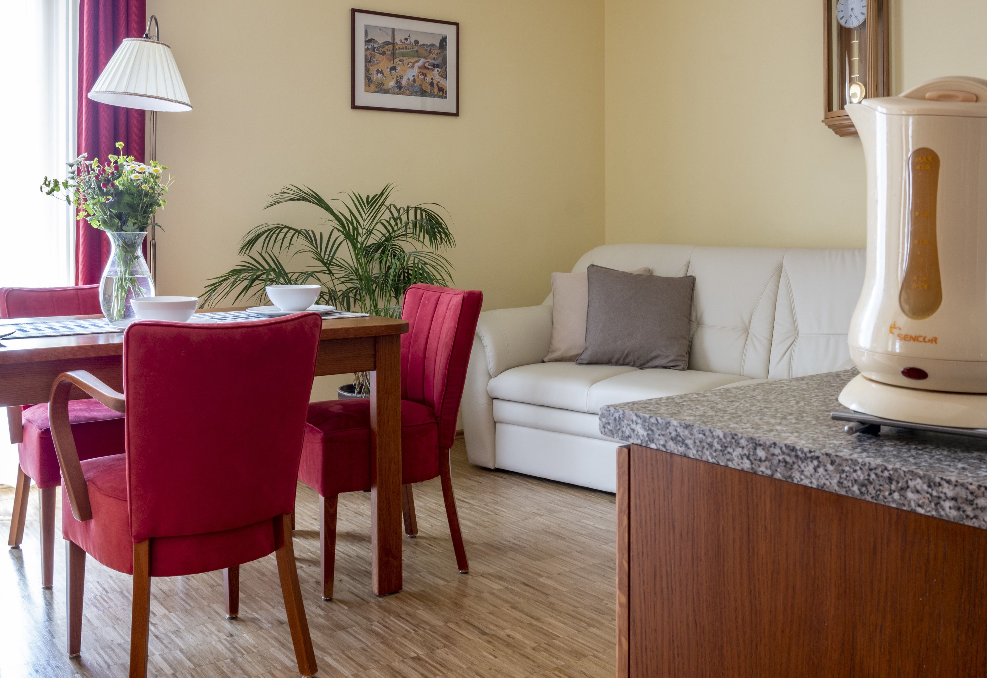 Een moderne woonkamer met een witte bank, rode stoelen, een eettafel met een bloevas, een potplant en een keuken met een koffiezetapparaat.