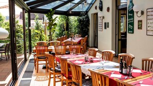 An outdoor dining area with tables and chairs, a glass roof, and a view of the surrounding greenery. - Hotel Pavilon (Ricany)