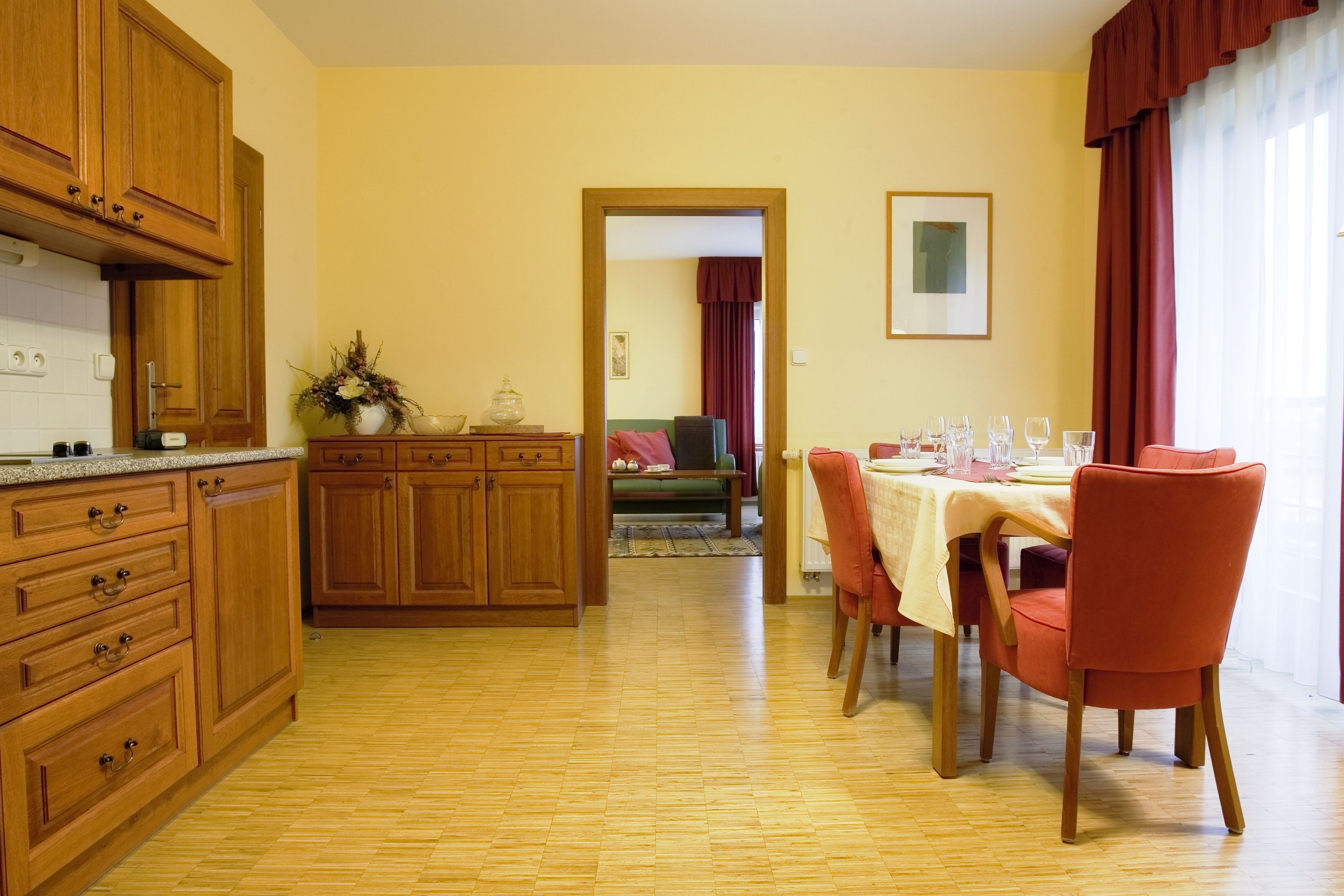Une cuisine avec des meubles de rangement en bois, une table à manger avec des chaises, et une vue sur le salon par une porte ouverte.