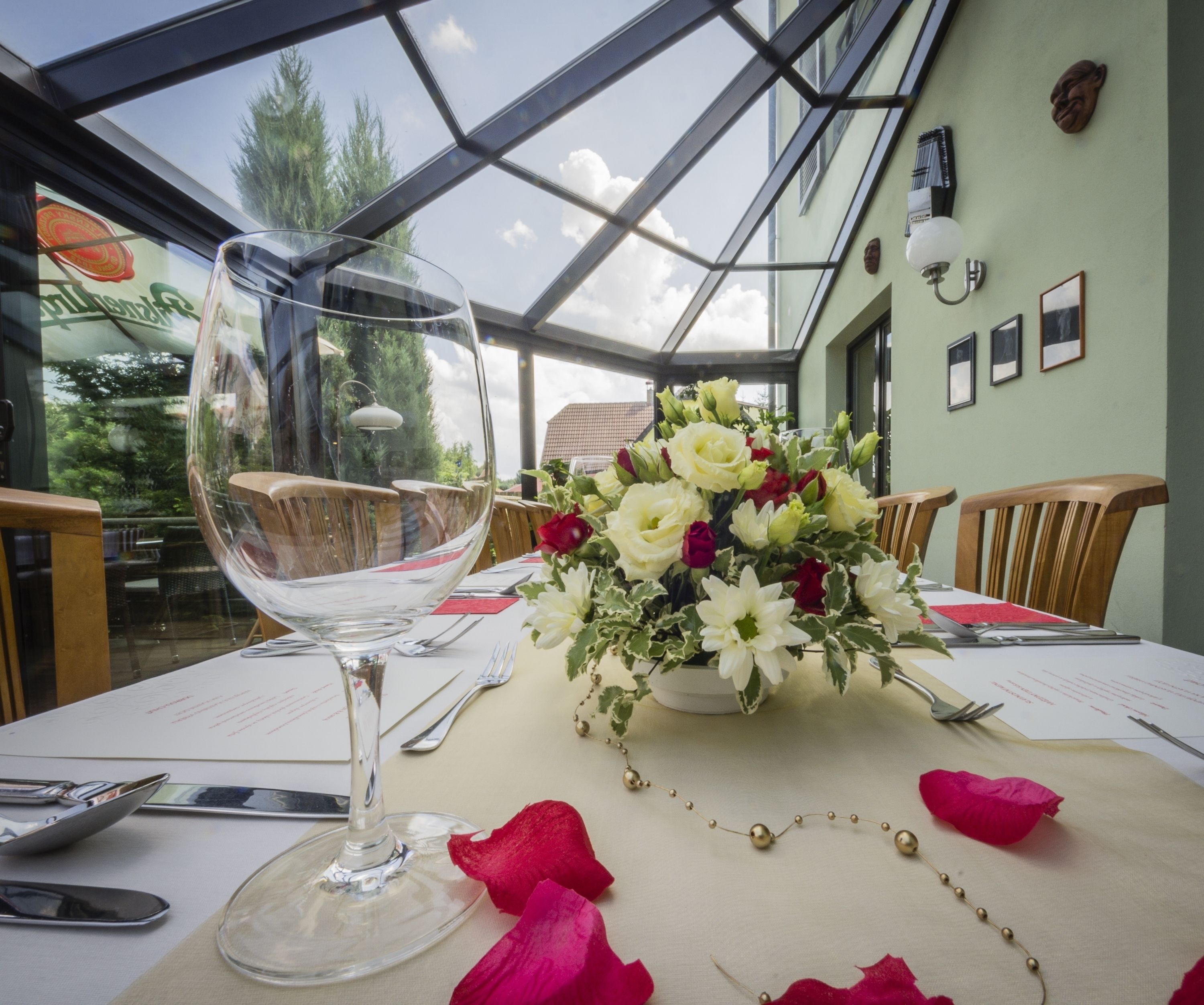 Mesa posta para refeição com um copo de vinho, um vaso de flores e pétalas de rosa sobre a toalha.