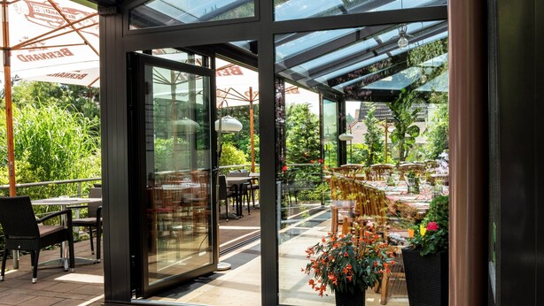 An indoor/outdoor dining area with glass walls, outdoor seating, and potted plants. - Hotel Pavilon (Ricany)
