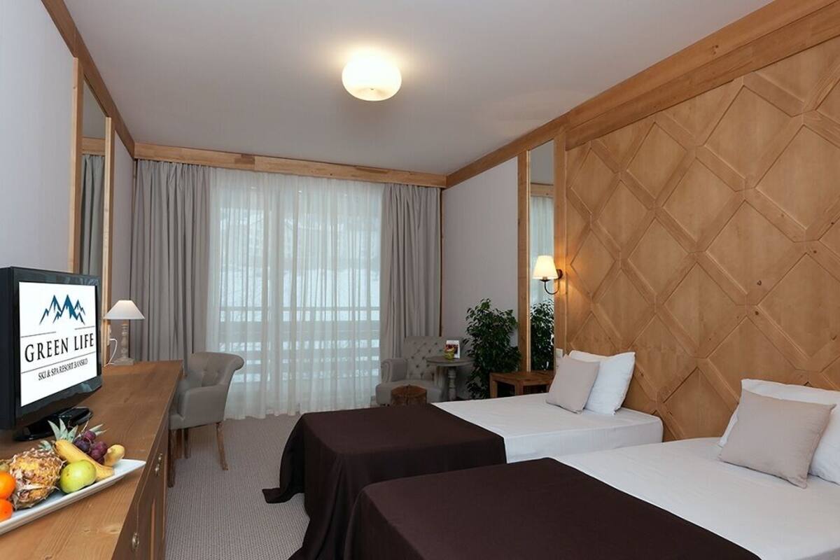 A hotel room with two beds, a TV, a desk with a fruit tray, and a large window with curtains.