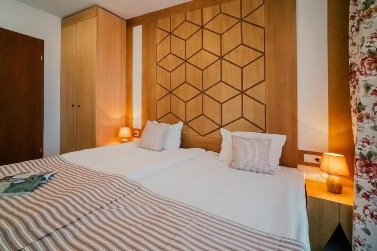 Hotellihuone, jossa on kaksi sänkyä, puinen pääty, yöpöytävalaisimet ja raidallinen päiväpeite.