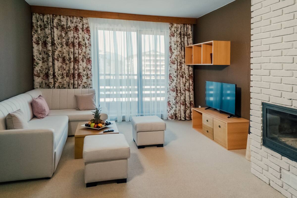 A modern living room with a sofa, ottomans, a coffee table with fruit, a TV, and a fireplace.