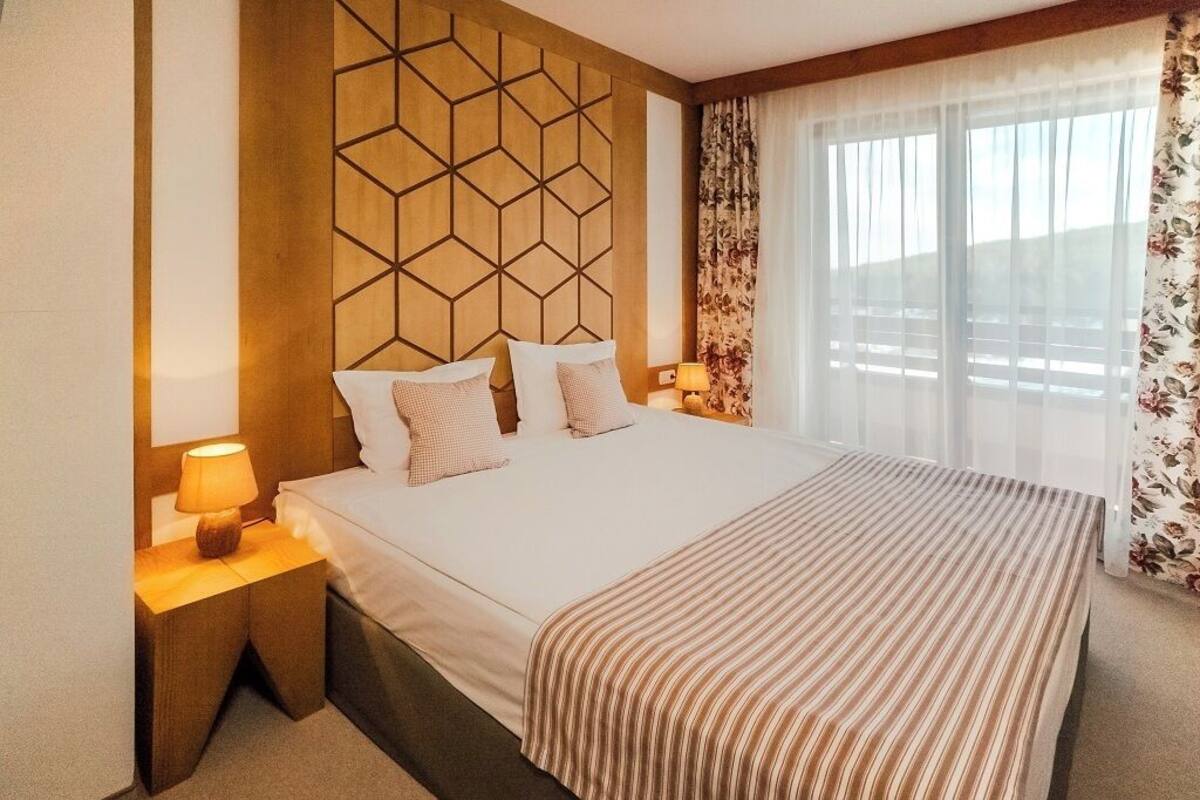 A hotel room with a large bed, wooden headboard, two bedside tables with lamps, and a window with sheer curtains.