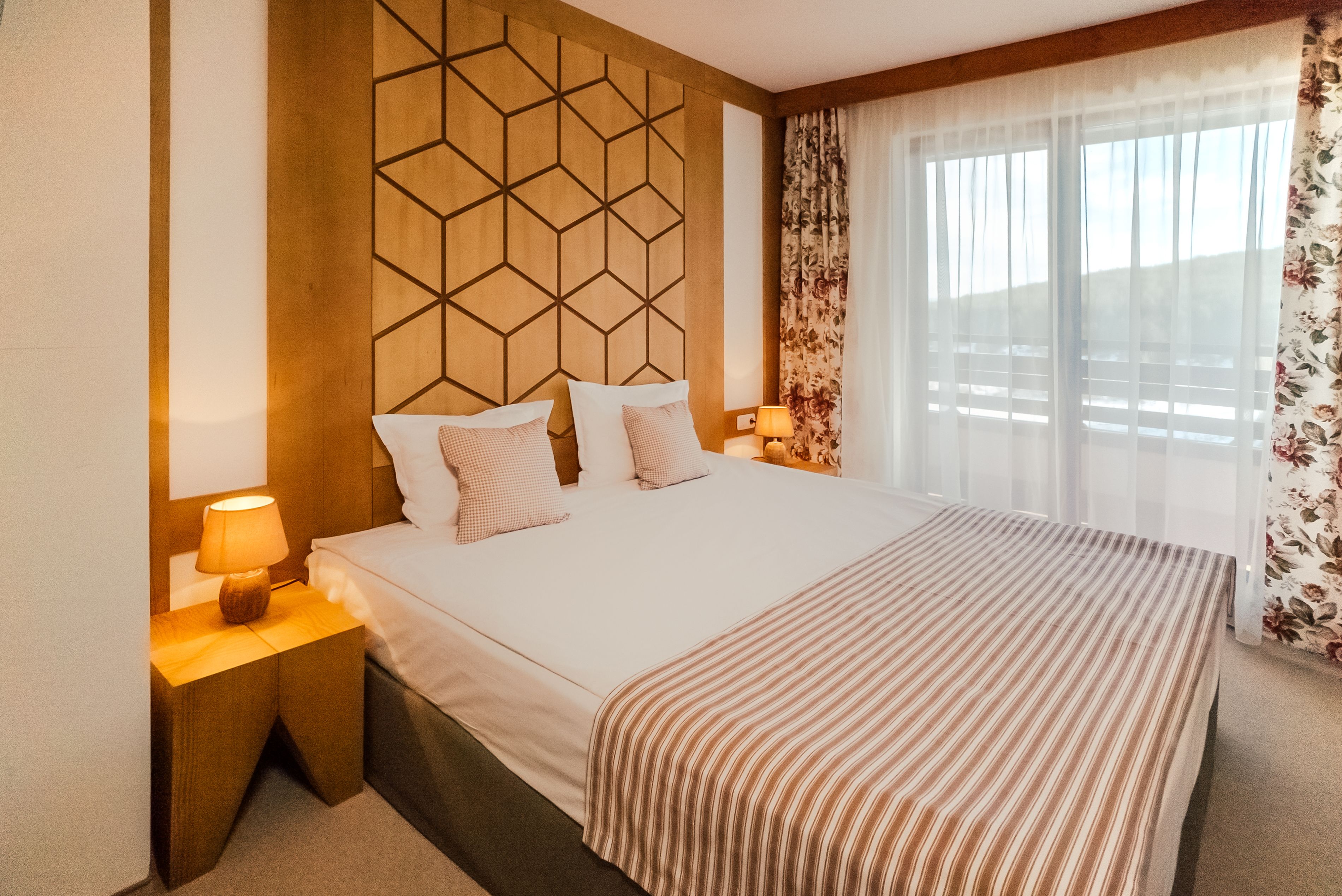 Habitación de hotel con una cama grande, cabecero de madera, dos mesitas de noche con lámparas y vistas a través de cortinas de gasa.