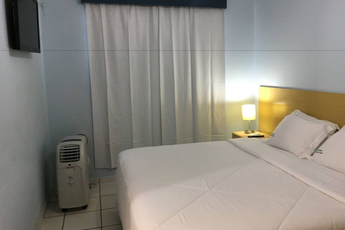 A hotel room with a bed, a nightstand with a lamp, a wall-mounted TV, and a window with curtains.