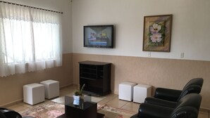 Una sala de estar con un televisor de pantalla plana, un cuadro floral y una planta en maceta sobre una mesa de centro de vidrio.