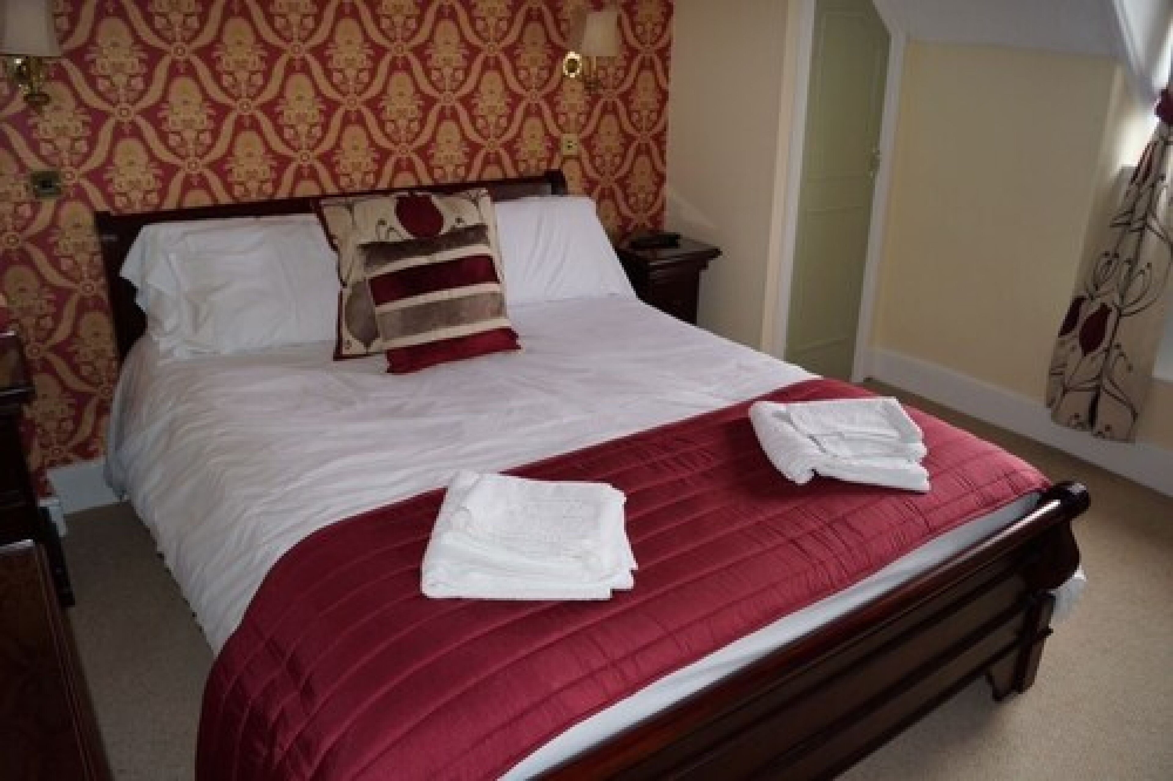 A neatly made bed with a red bedspread and white linens, a wooden headboard, and a patterned wallpaper.