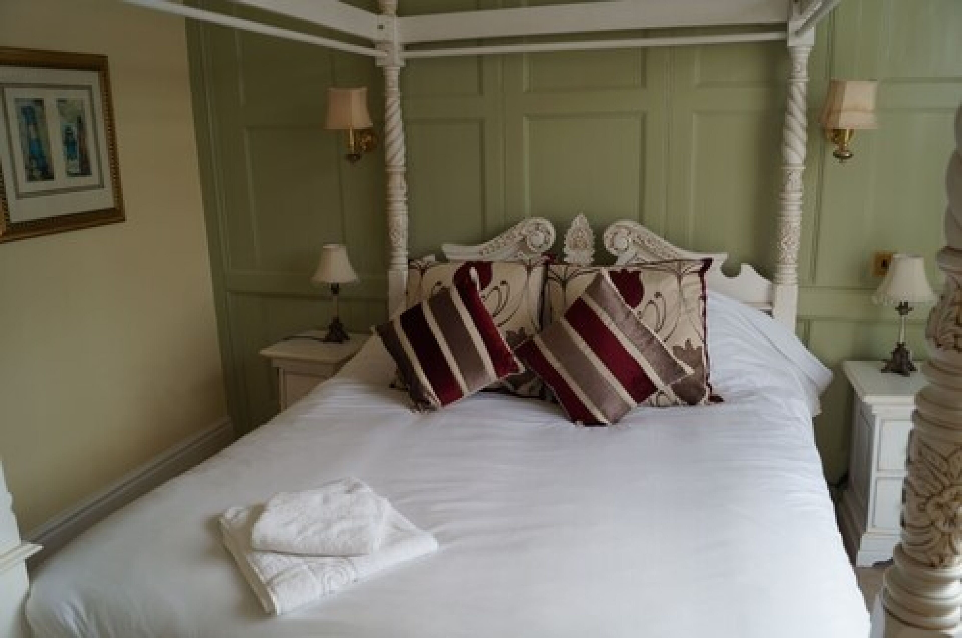 A four-poster bed with white bedding and striped pillows, a white nightstand with a lamp, and a framed picture on the wall.