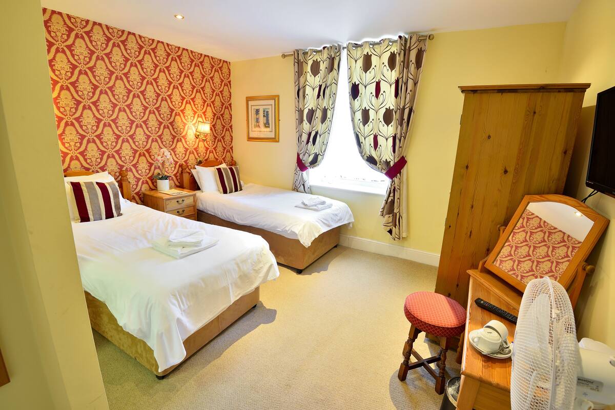 A hotel room with two beds, a wooden wardrobe, a desk with a chair, a television, and a window with curtains.