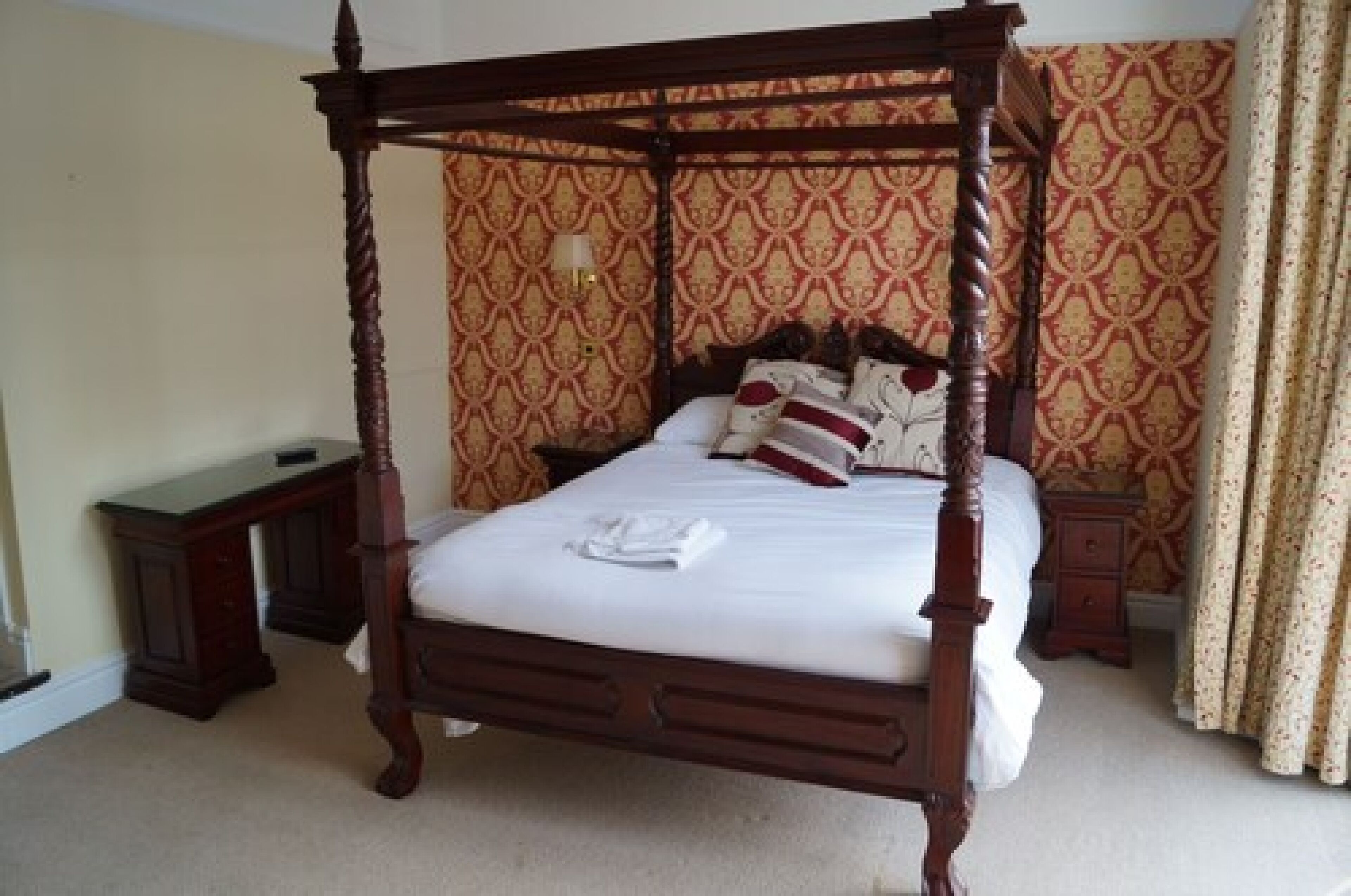 A four-poster bed with a wooden frame, a bedside table, a nightstand, and a patterned wallpaper.