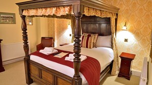 A four-poster bed with a red bedspread and matching pillows, a nightstand with a lamp, and a wall-mounted picture frame. - The Angel Hotel (Coleford)
