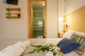 A hotel room with a bed, a wooden headboard, a nightstand, a wall-mounted TV, and a shower area visible through an open door. - The Nomad Hostel (Seville)