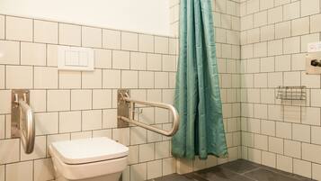 Une salle de bain avec des carreaux blancs, des toilettes blanches, une barre d’appui, un rideau de douche turquoise et un sèche-mains.