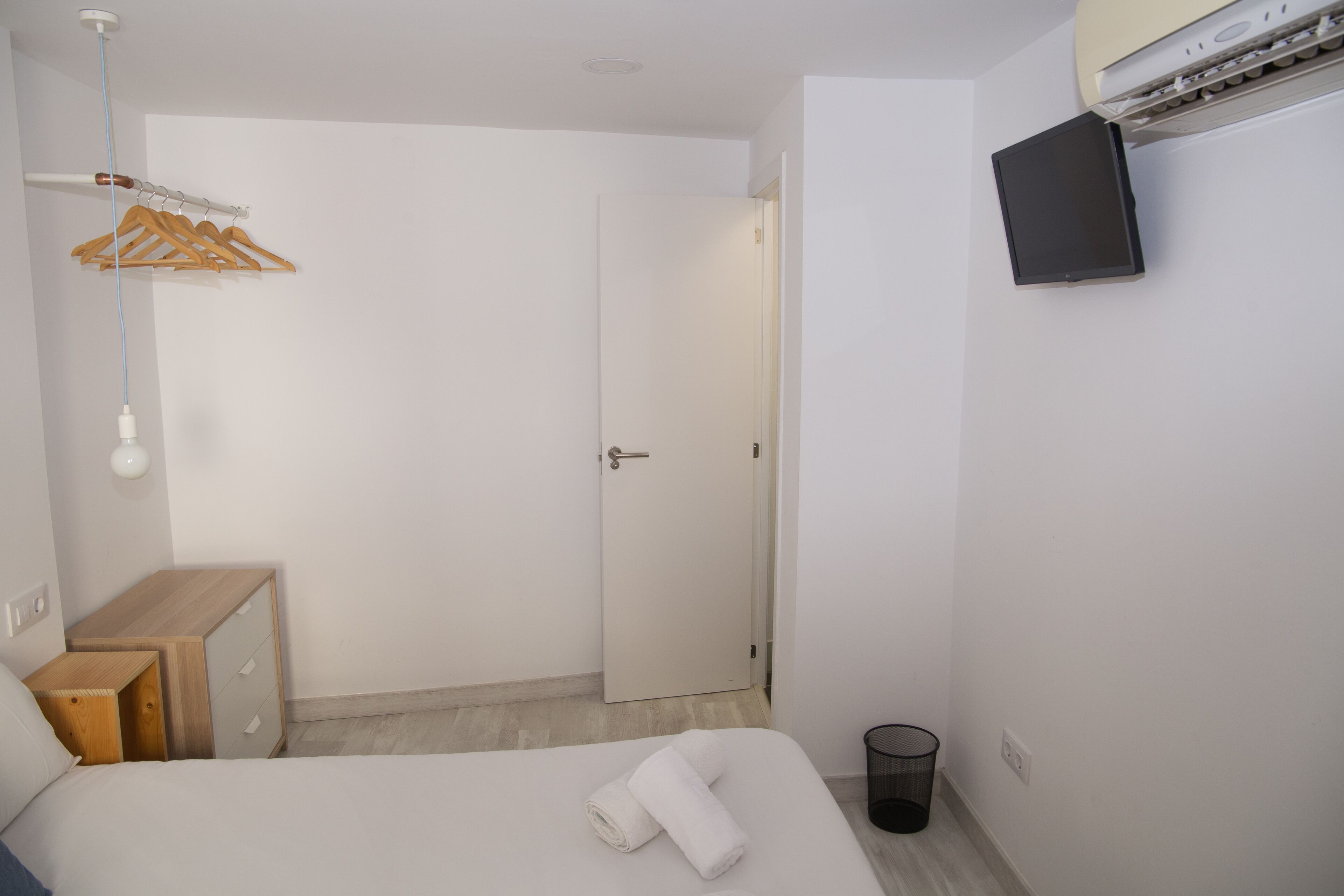 A hotel room with a bed, a wooden nightstand, a wall-mounted TV, a door, and a hanging coat rack.