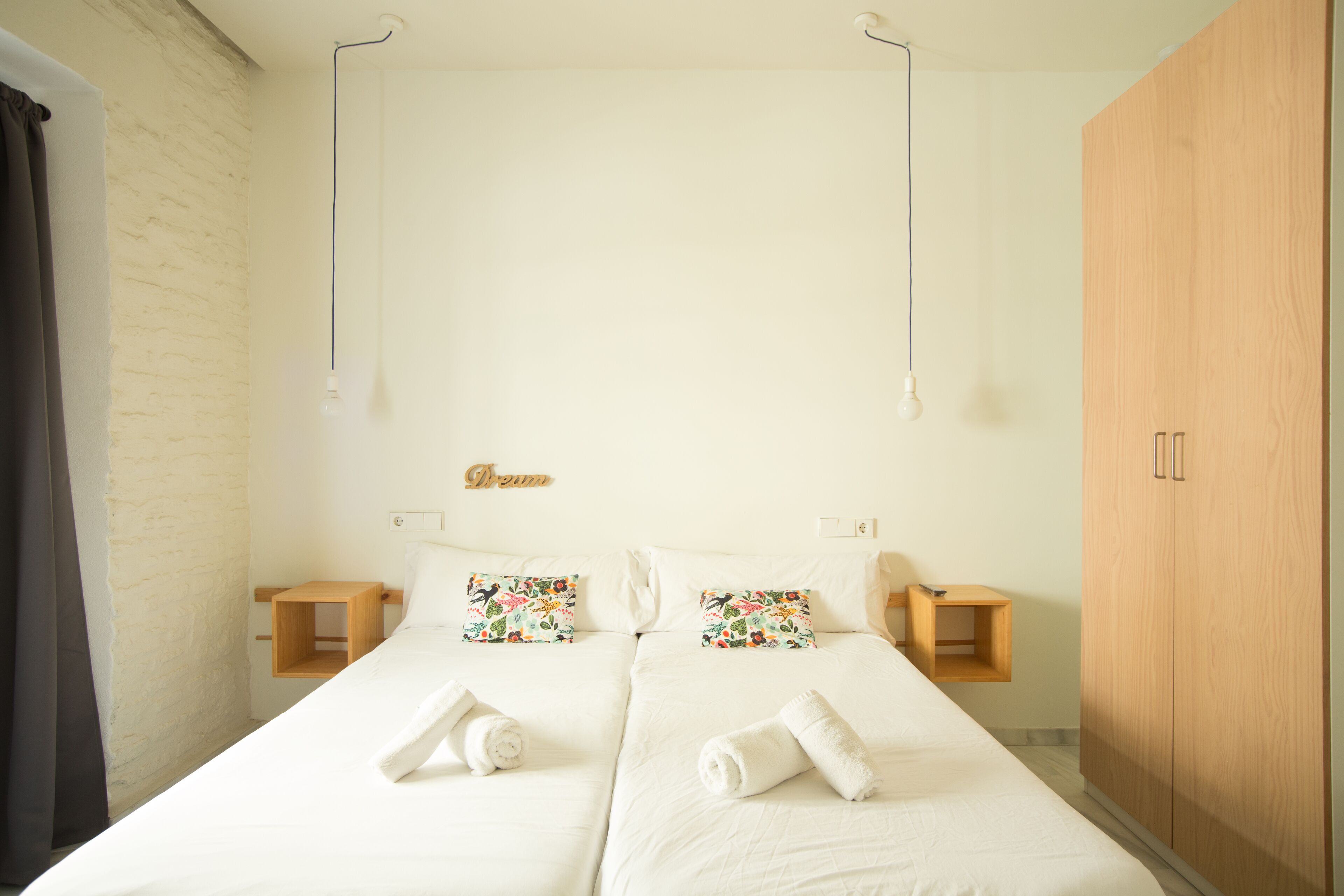 A double bed with floral pillows, a wooden nightstand, a wooden wardrobe, and a light fixture.
