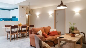 A modern living room with a sofa, dining table, and chairs. There is a kitchen area in the background with blue tiles. - The Nomad Hostel (Seville)