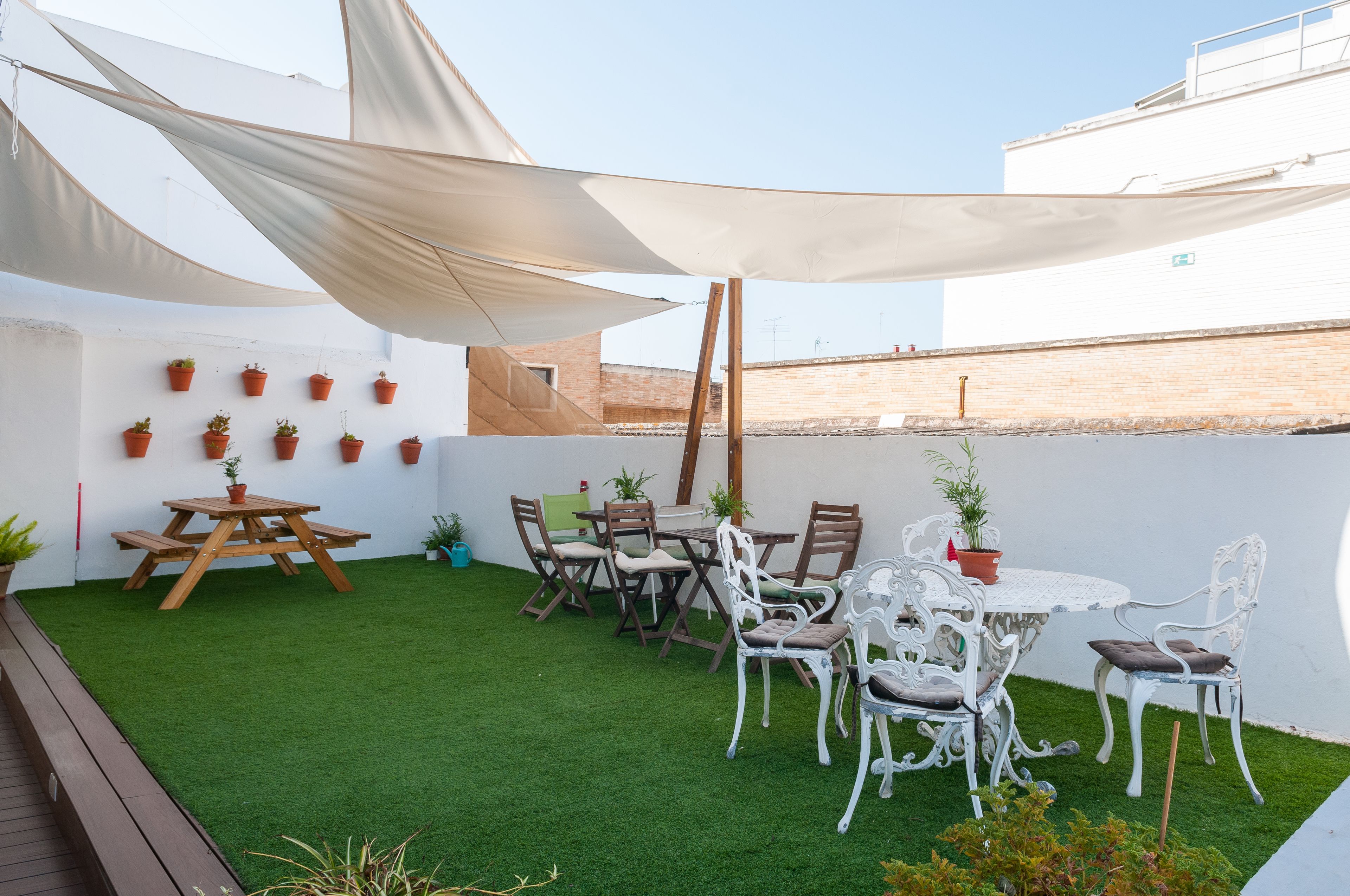 Un patio con una mesa y sillas, rodeado de plantas en macetas y un muro blanco.