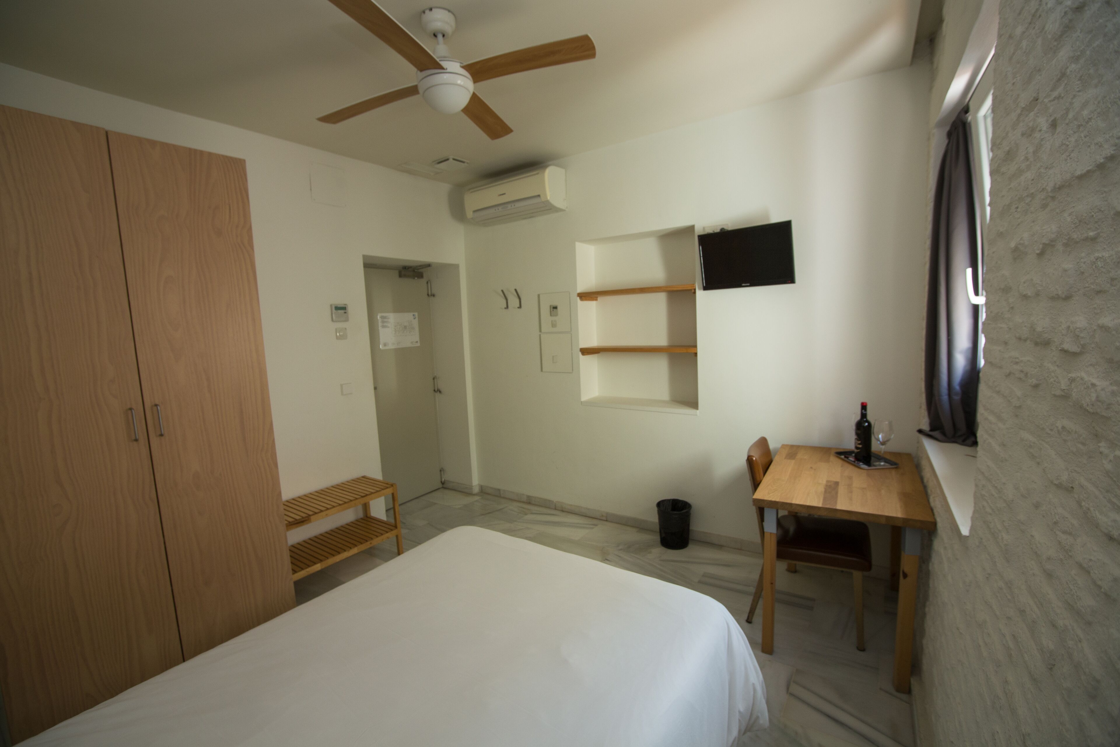 A bedroom with a bed, wooden wardrobe, ceiling fan, air conditioning unit, and a small table with a chair.