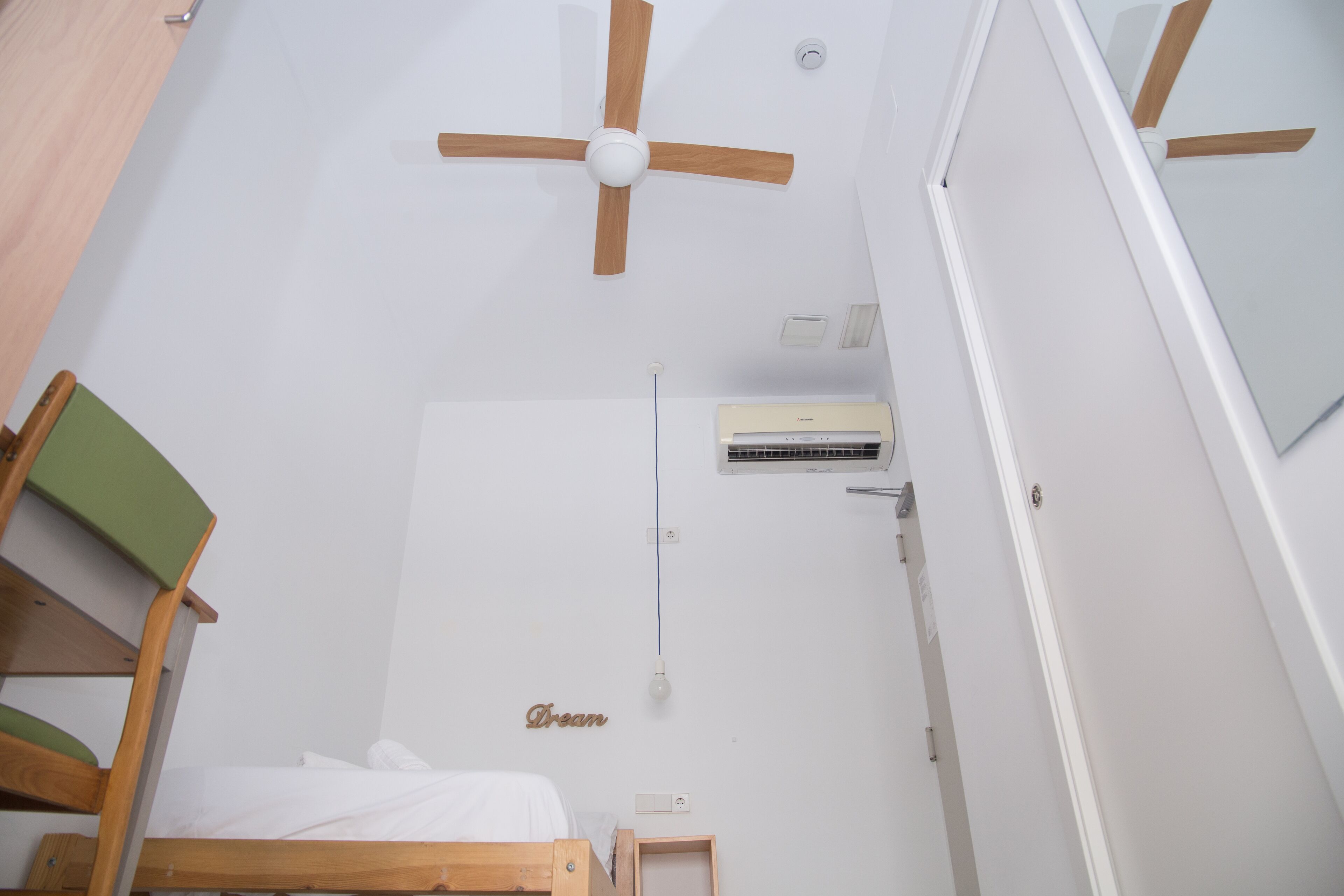 A small, white room with a single bed, a ceiling fan, an air conditioning unit, and a wooden chair.