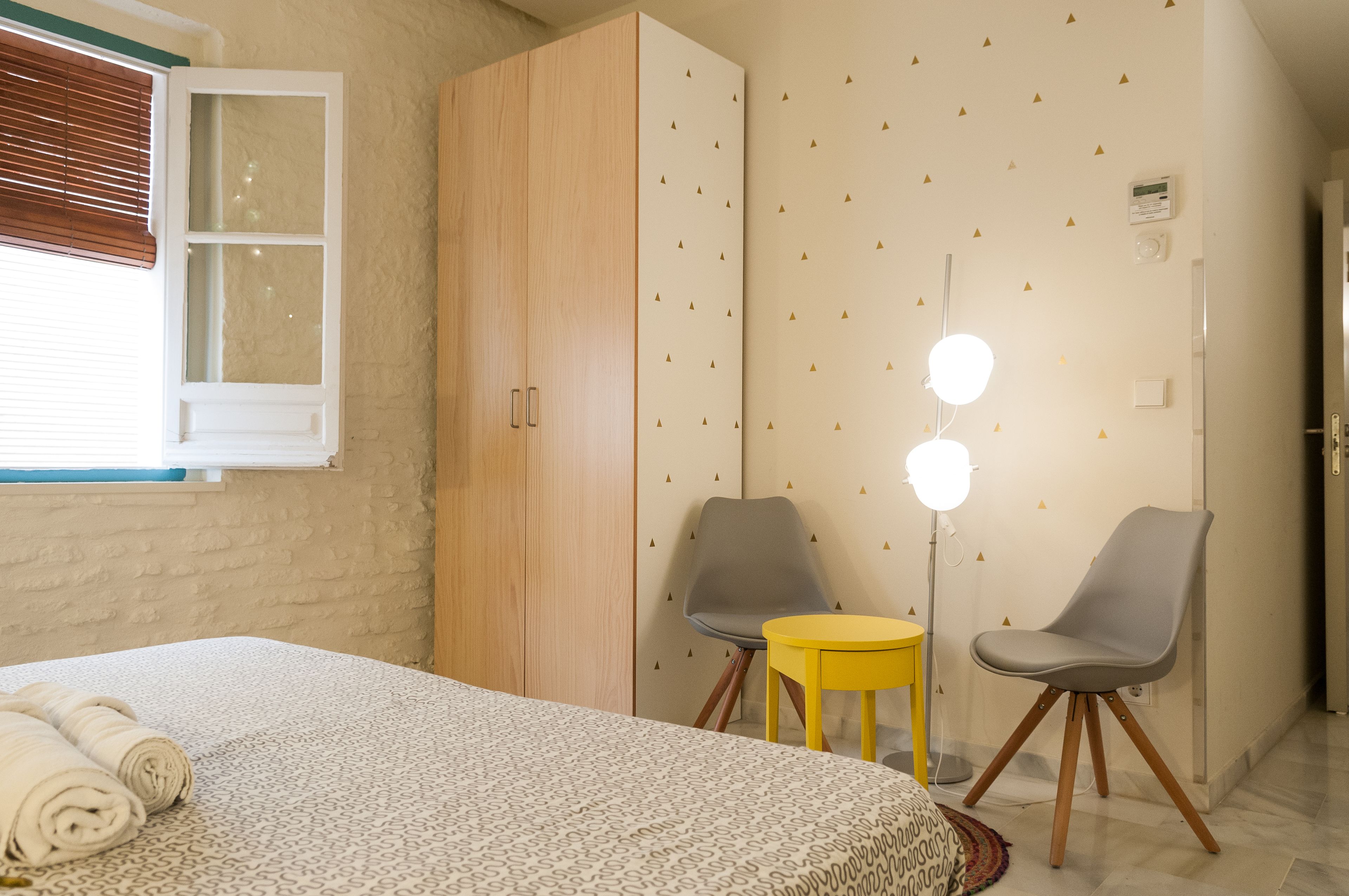 A bedroom with a bed, wardrobe, two chairs, a small table, and a wall-mounted light fixture.