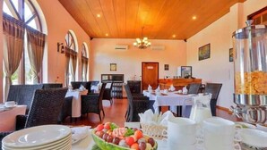Un coin repas avec des tables préparées pour le petit-déjeuner, comprenant un panier de fruits et des tasses à café.