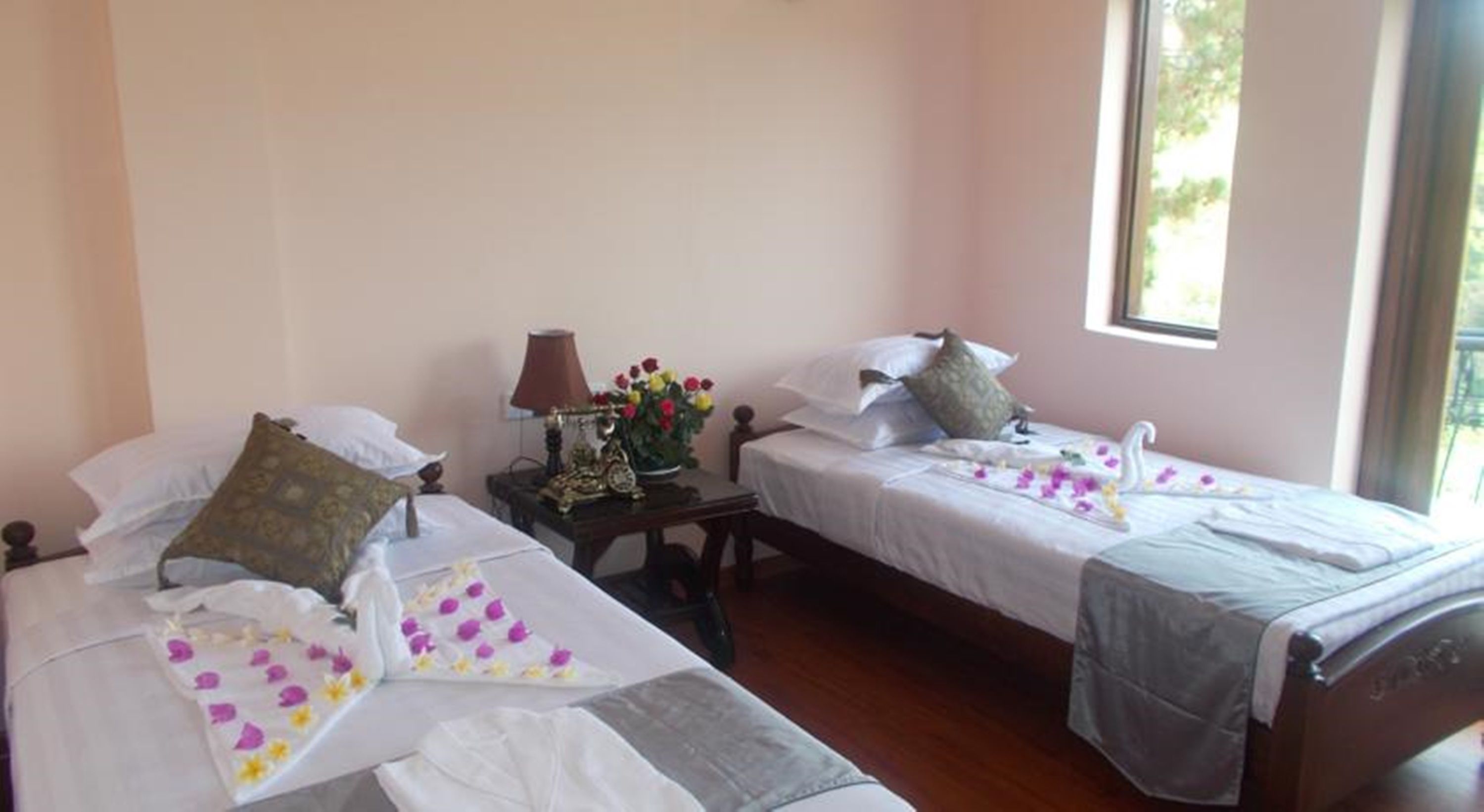 A room with two single beds, each with a pillow and a bedside table with a lamp and a vase of flowers.