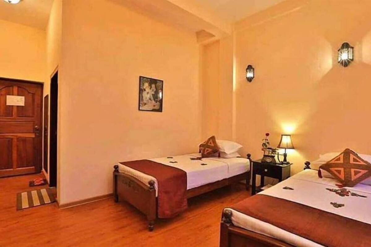 A hotel room with two single beds, wooden flooring, a door, a small bedside table with a lamp, and a wall-mounted light fixture.