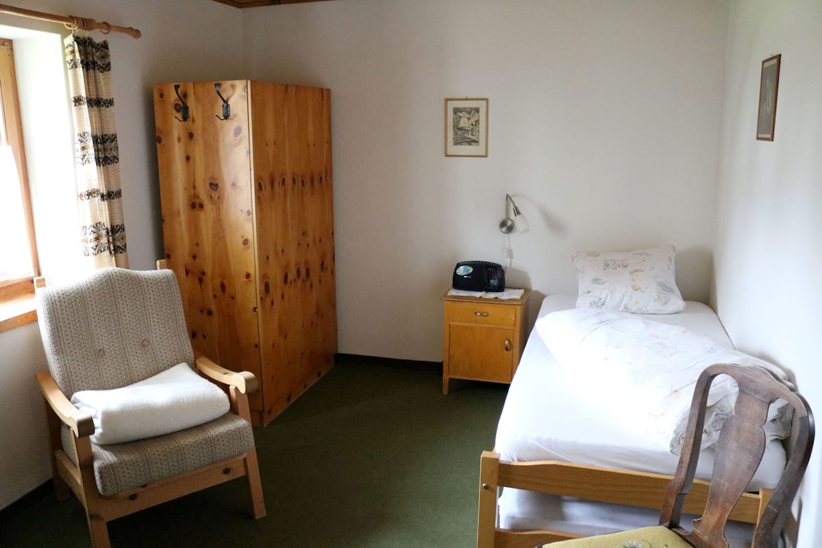 A bedroom with a wooden bed, a wooden chair, a nightstand with a radio, and a wardrobe.