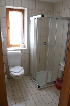 Un baño pequeño con inodoro, ventana con cortina, cabina de ducha con mampara de vidrio y un cubo de basura rojo.