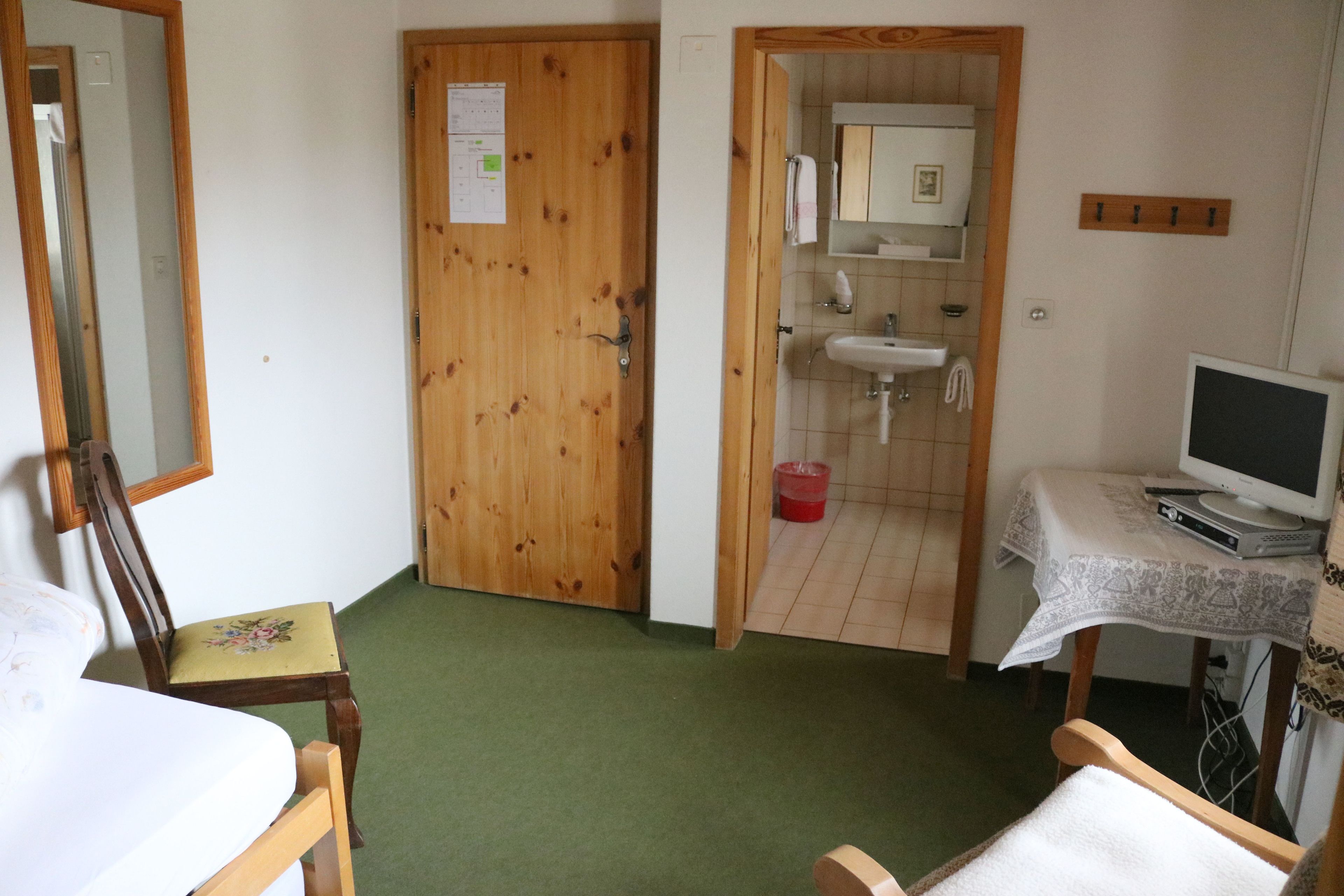 A room with a wooden door, a bed, a chair, a desk with a computer, and a bathroom visible through an open door.