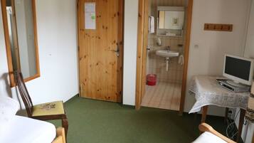 A room with a wooden door, a bed, a chair, a desk with a computer, and a bathroom visible through an open door.
