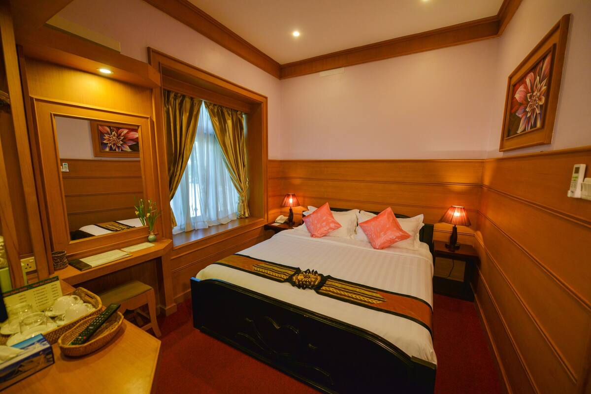 A hotel room with a large bed, bedside tables, a desk with a chair, a window with curtains, and a painting on the wall.