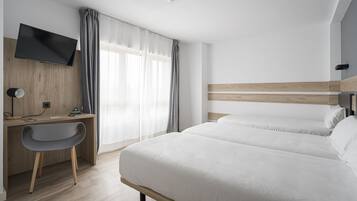 Habitación de hotel con cama, escritorio con silla, televisor montado en la pared y ventana con cortinas.