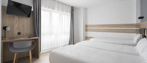 A hotel room with a bed, a desk with a chair, a TV mounted on the wall, and a window with curtains.