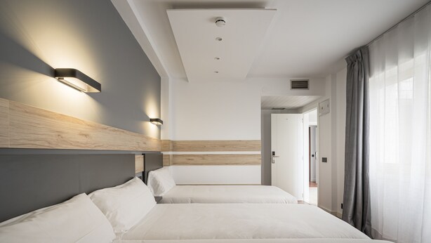 A hotel room with two beds, a wooden headboard, a light fixture, and a view of a hallway. - Alda Centro Gijón (Gijon)