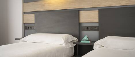 A modern hotel room with two beds, a nightstand with a lamp, and a wall-mounted control panel.