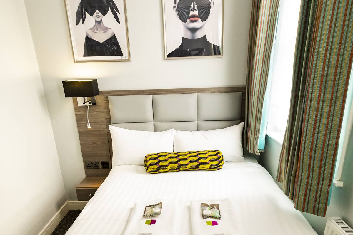 A hotel room with a large bed, a headboard, two framed artworks on the wall, a bedside lamp, and a window with curtains.