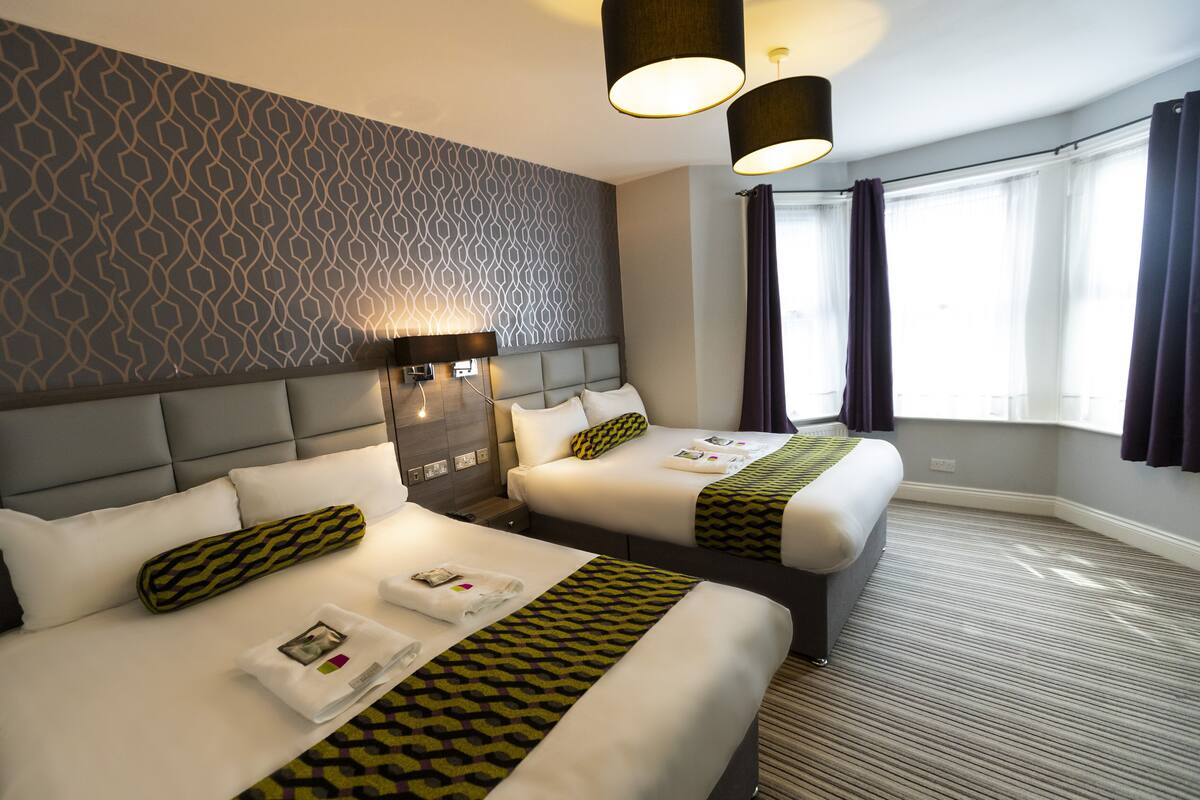 A hotel room with two beds, a patterned wall, a large window with curtains, and a modern light fixture.