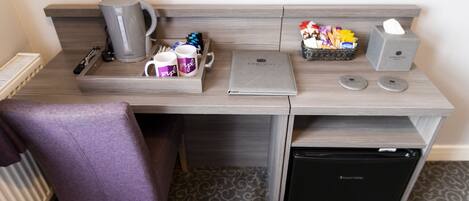 A hotel room with a desk, a purple chair, a coffee maker, a laptop, a tissue box, and a mini fridge.