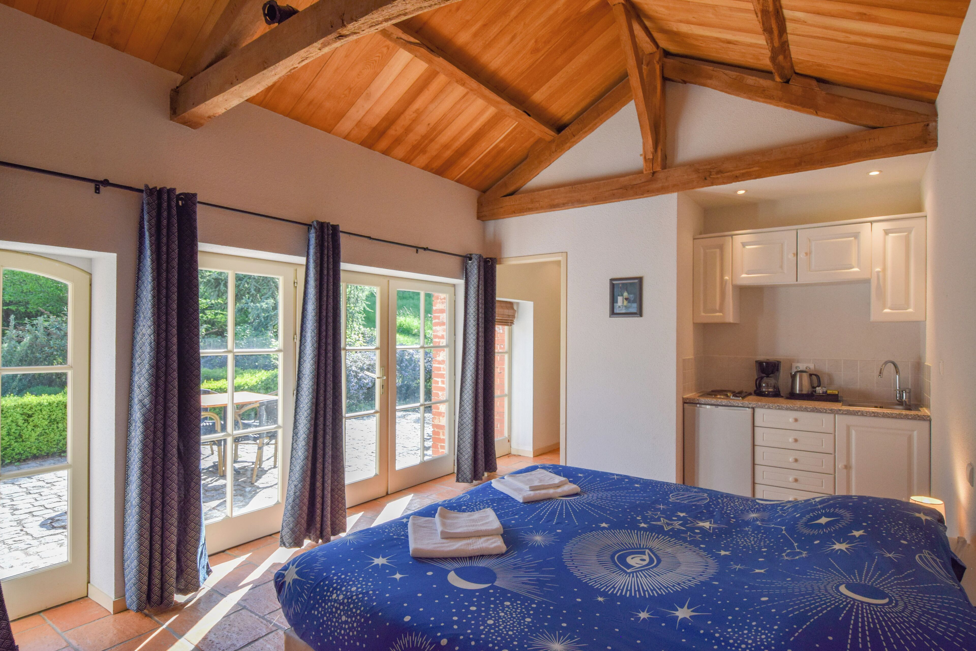 A bedroom with a large bed, a view of the garden through the window, and a kitchenette in the corner.