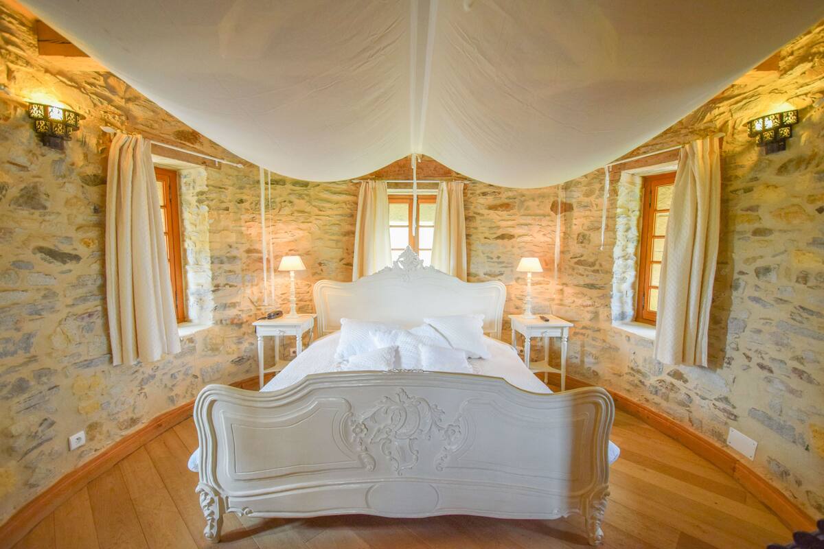 Un dormitorio con pared de piedra, cama blanca con dosel y cabecero ornamentado, y dos mesitas de noche con lámparas.