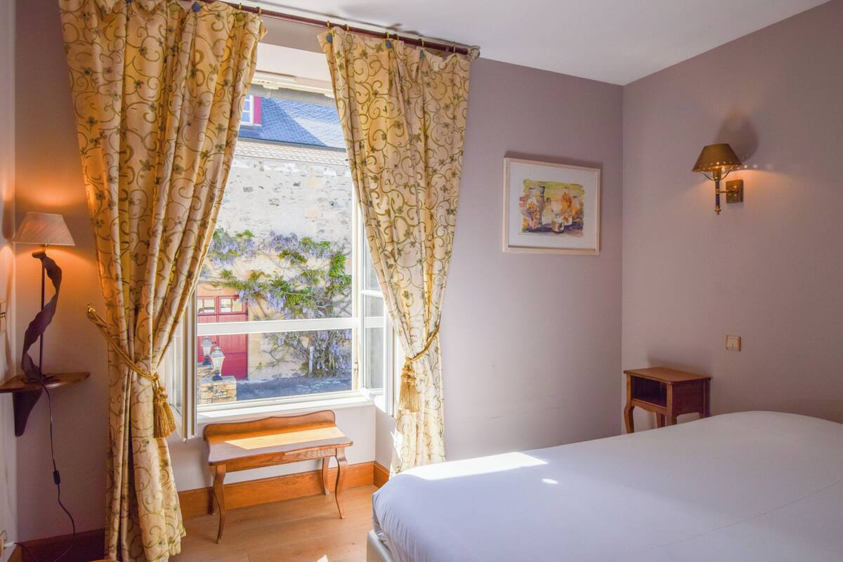 Un dormitorio con cama, ventana con cortinas, una mesita, una lámpara y una lámpara de pared.