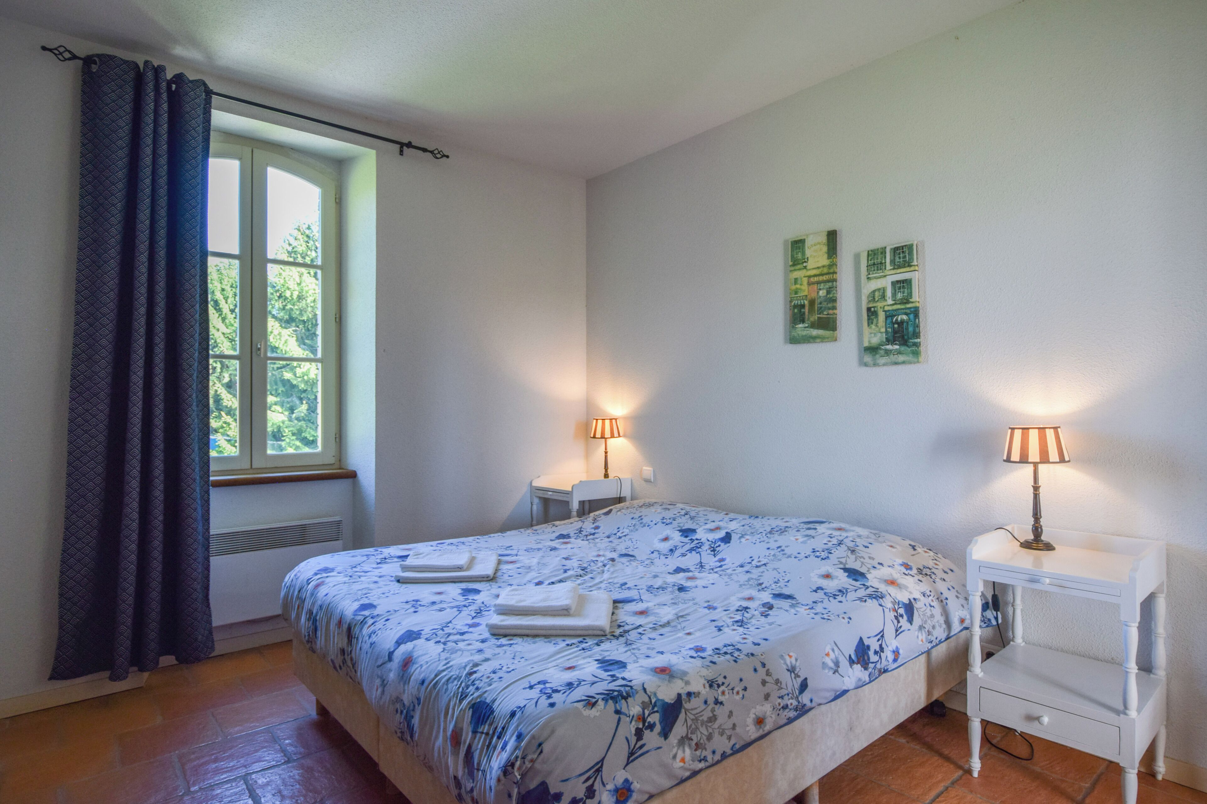 Un dormitorio con una cama, dos mesitas de noche, un ventanal con cortinas y dos lámparas de pared.